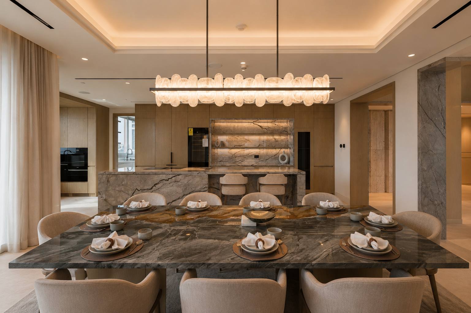 Dining room with grey-veined marble table, alabaster linear chandelier and open kitchen behind