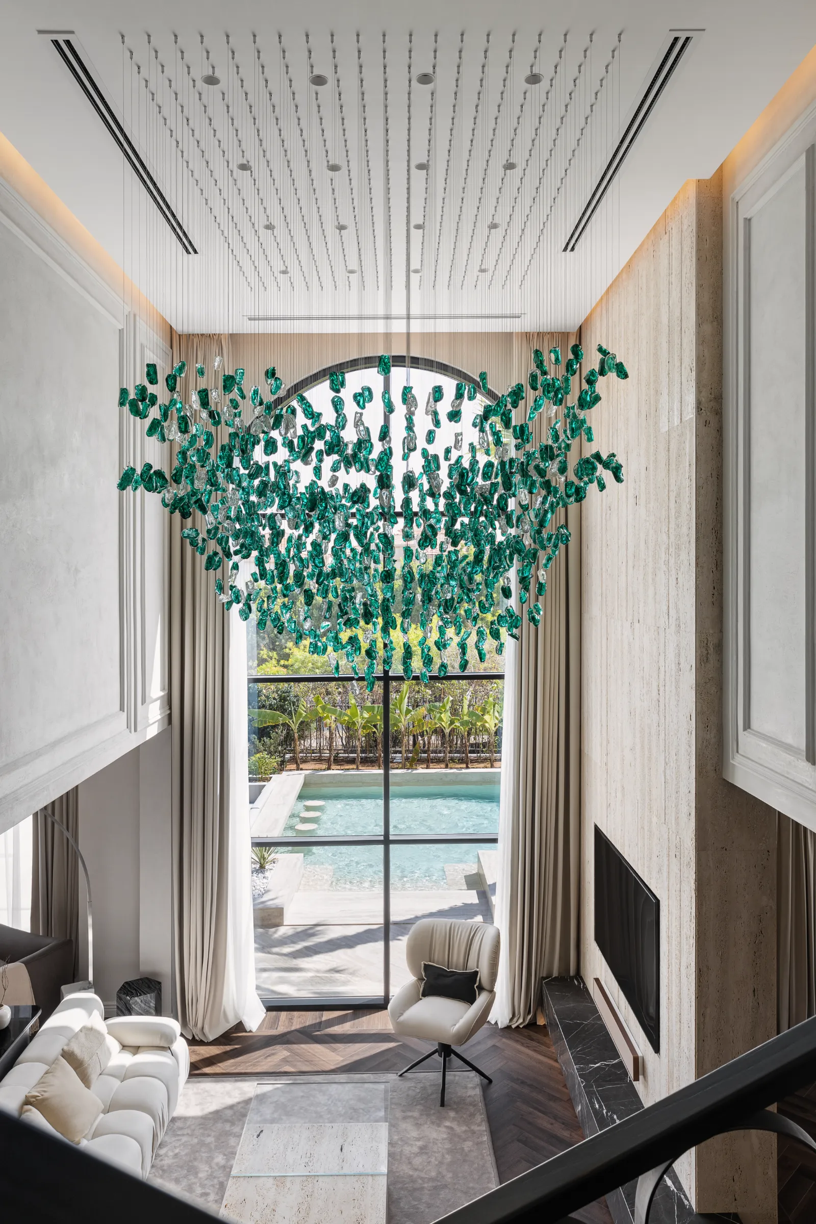 Double-height living room with cascading emerald glass chandelier and arched window to the pool