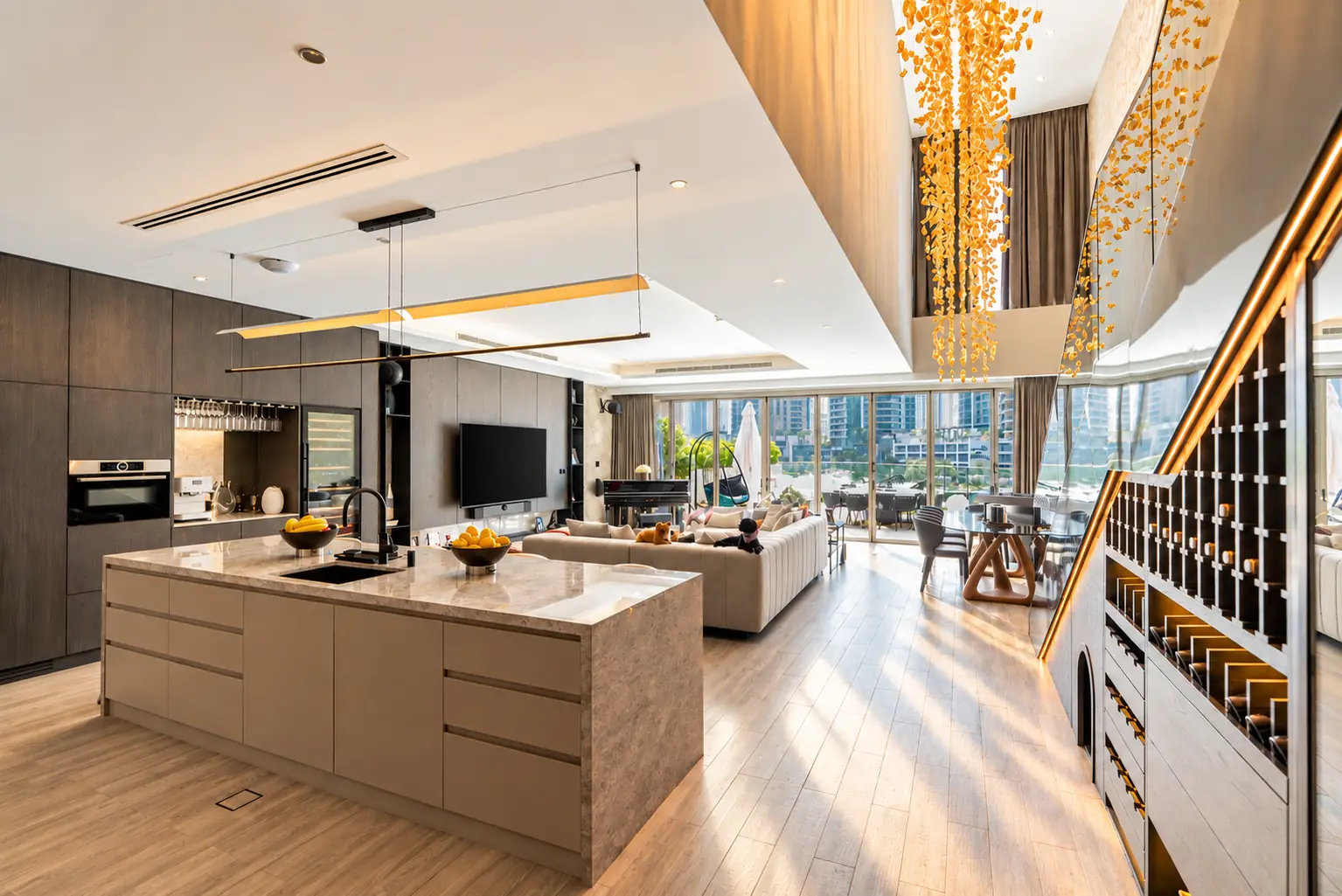 Open-plan kitchen and living room under a cascading amber glass chandelier in a double-height hall, with folding glass doors to a marina terrace, Villa Ambra by K4