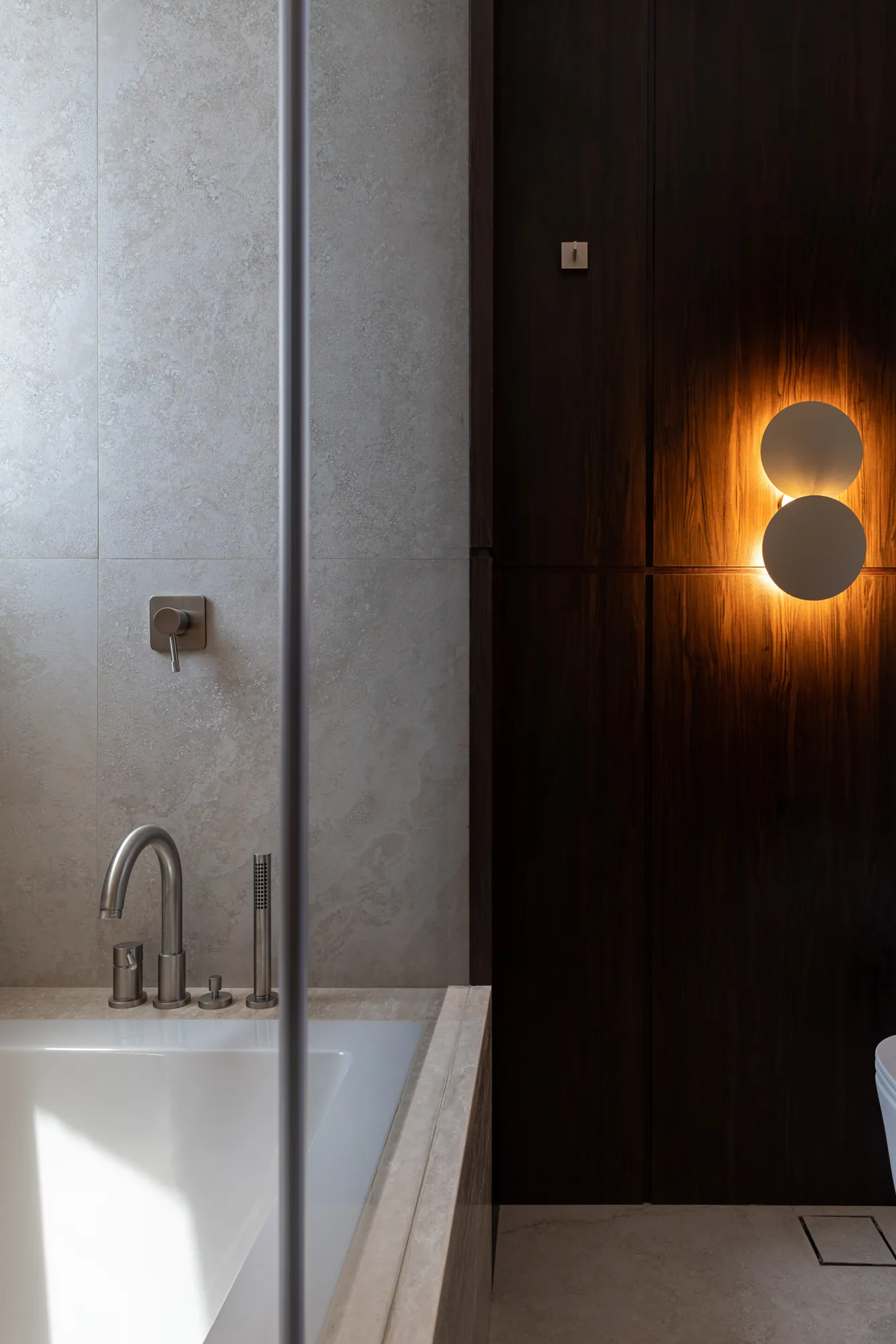 Bathtub with brushed fittings against stone tiles beside dark timber panelling and a glowing disc wall light