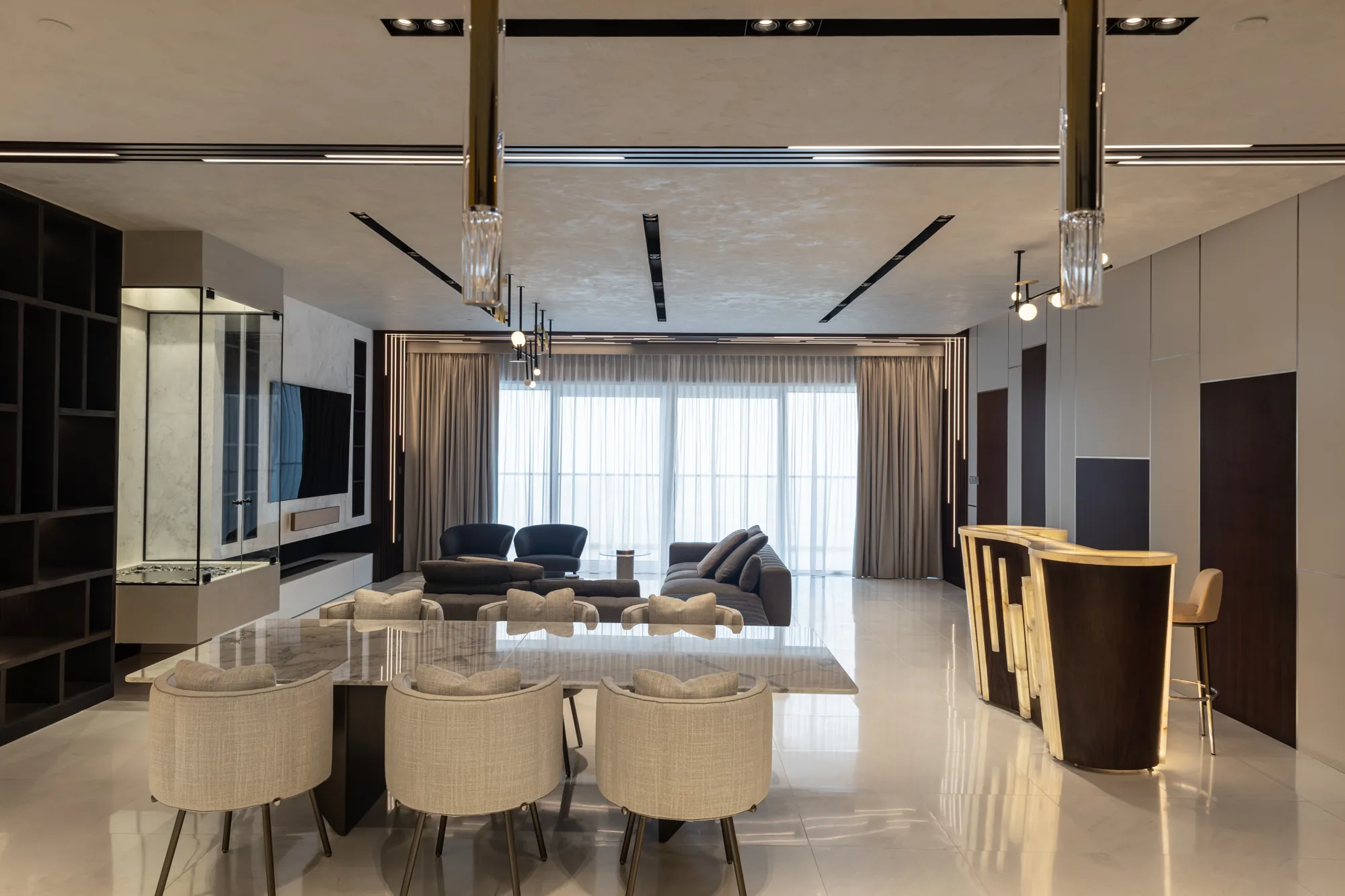 Open-plan dining and living room with a marble dining table, backlit onyx and walnut bar and sheer-curtained glazing, One JBR Apartment by K4