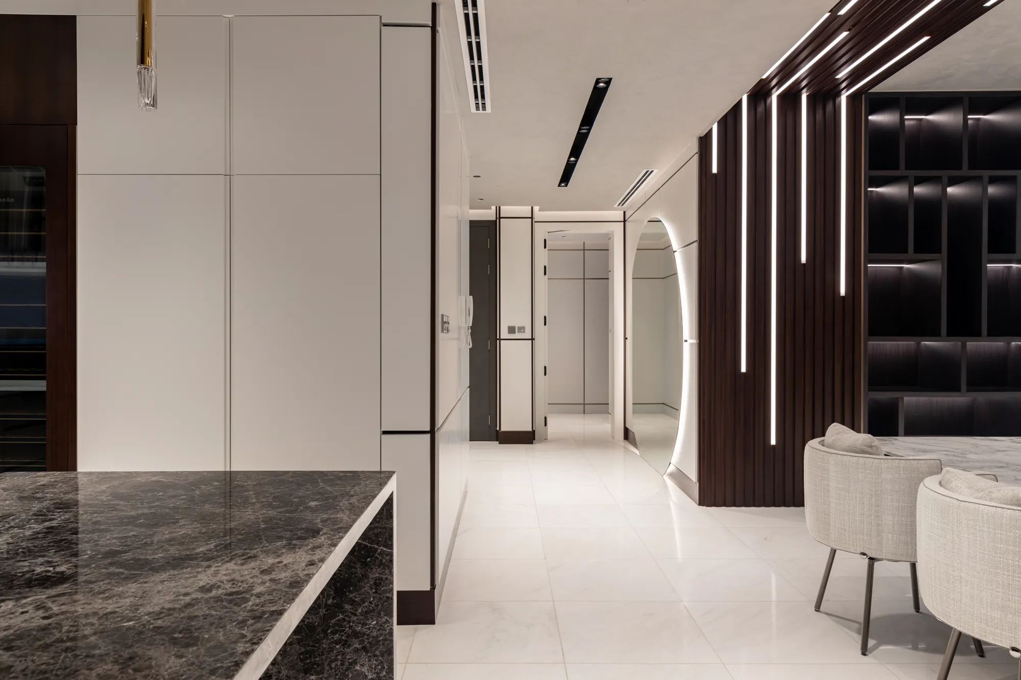 Dark marble kitchen island beside white handleless tall cabinetry, looking down a white gallery corridor past a backlit oval mirror and walnut slats