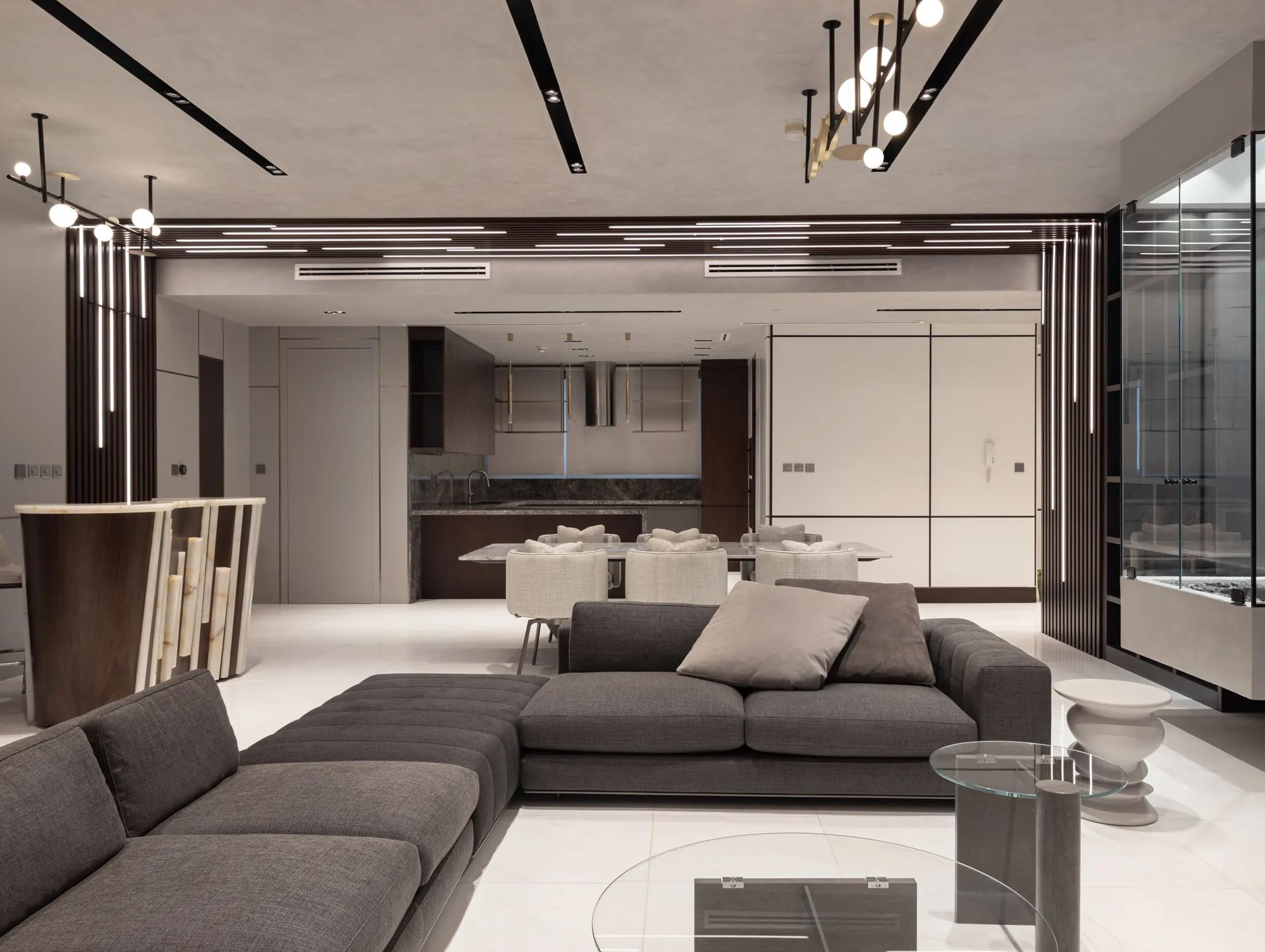 Living room with a grey modular sofa, glass coffee tables, a slatted walnut frame with linear lighting and the dining area and kitchen beyond